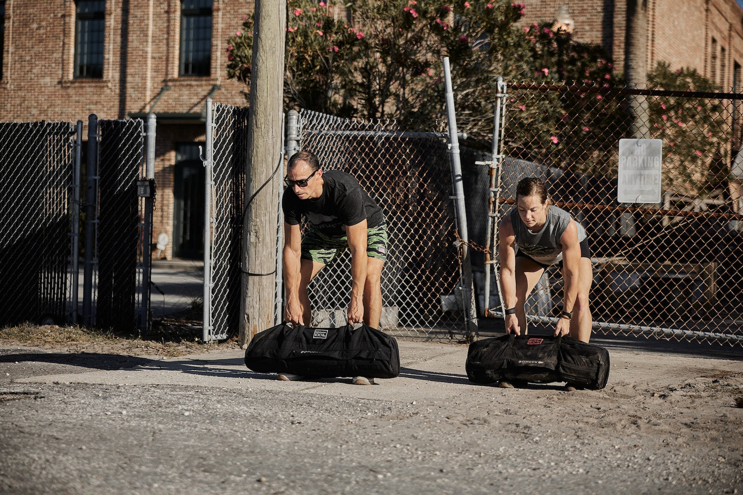 Two people outdoors lifting GORUCK sandbags in front of a chain link fence