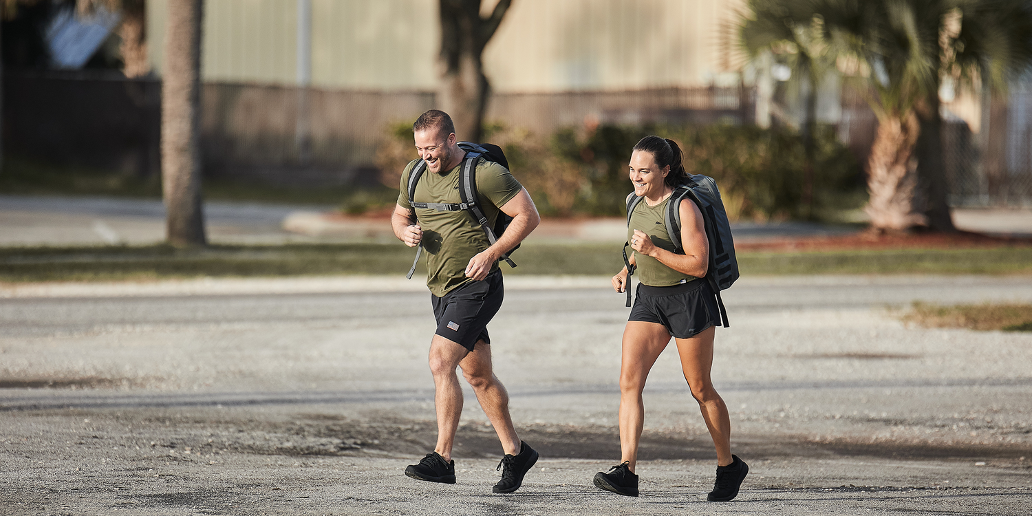Two people rucking outdoors with GORUCK backpacks and athletic gear in a sunny urban setting.