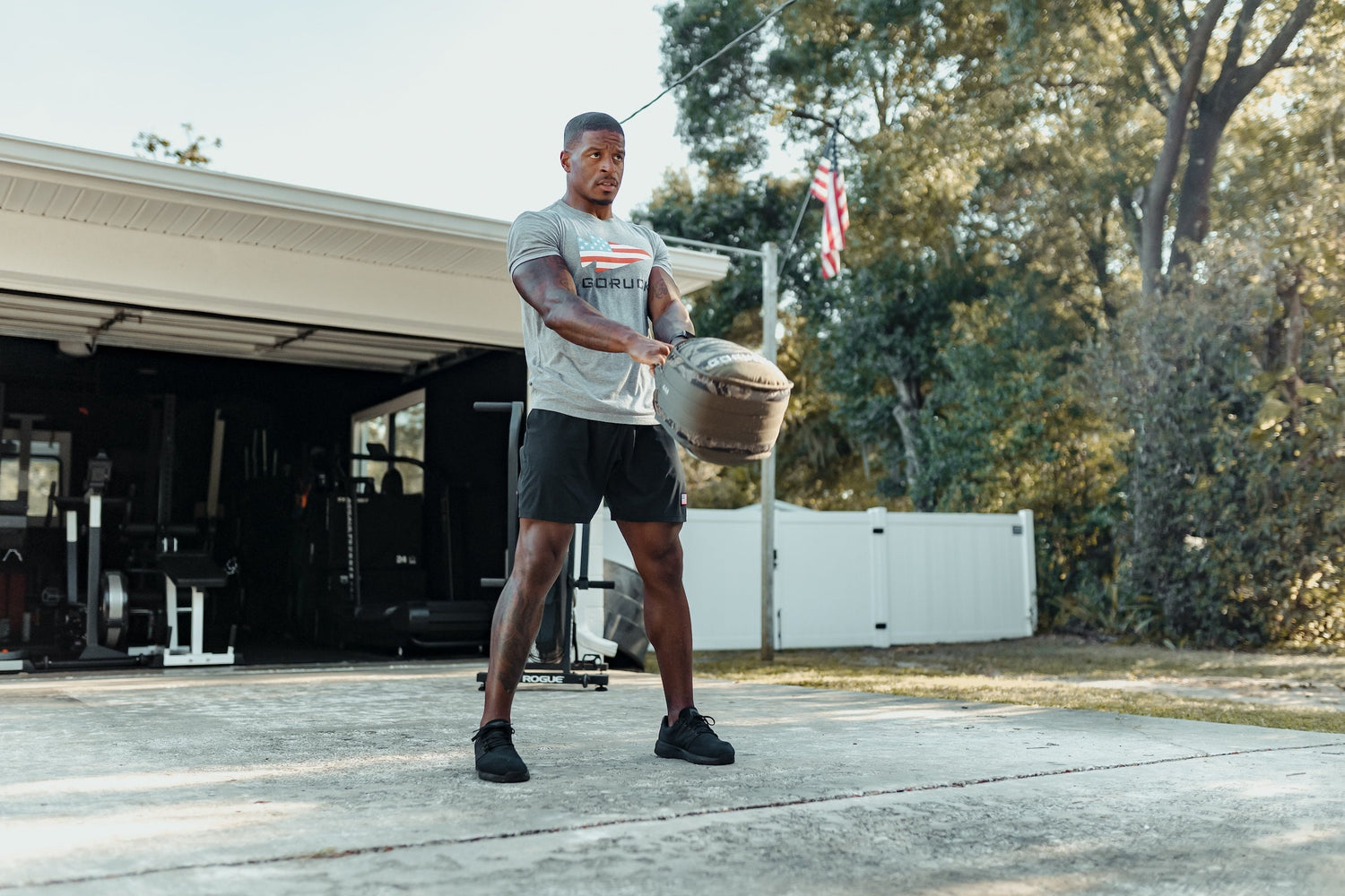 Man training outdoors with GORUCK sandbag, wearing branded GORUCK shirt and shorts.