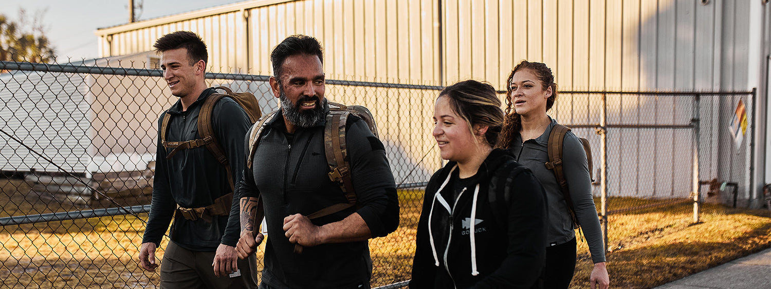 Group of people rucking with GORUCK backpacks outdoors near a fence in sunlight