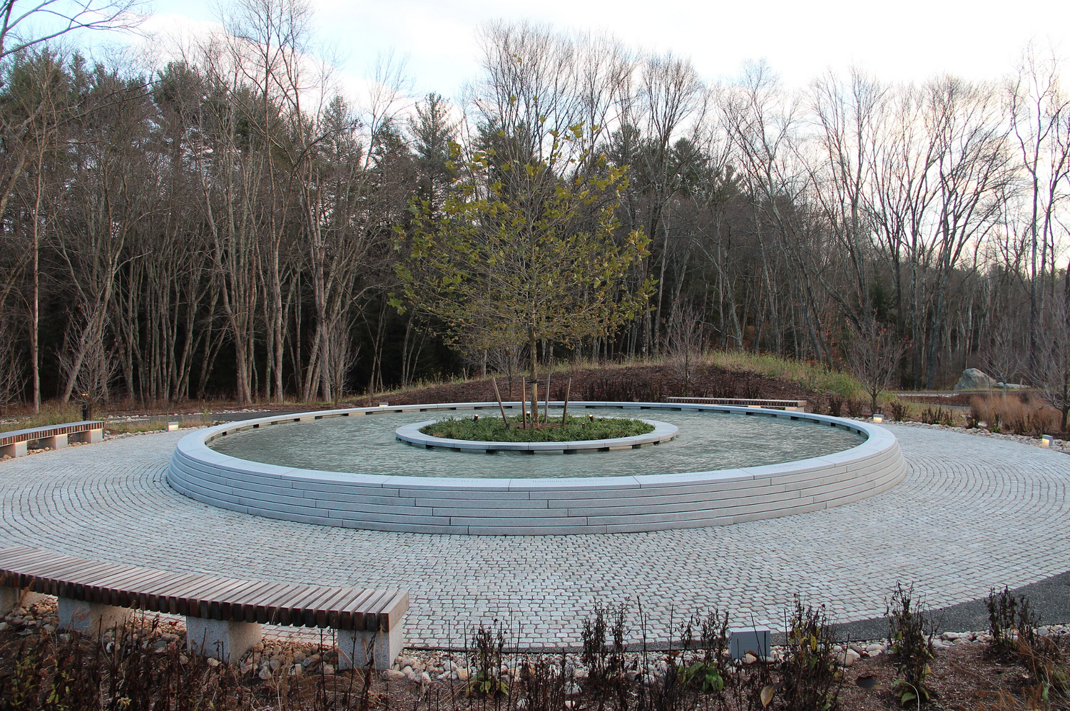 Circular memorial with central tree and reflecting pool, stone benches, and forest backdrop at GORUCK.