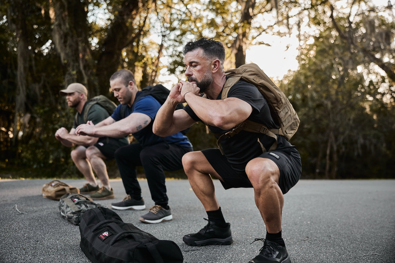 Three men training outdoors with GORUCK rucksacks, performing squats on a paved path.