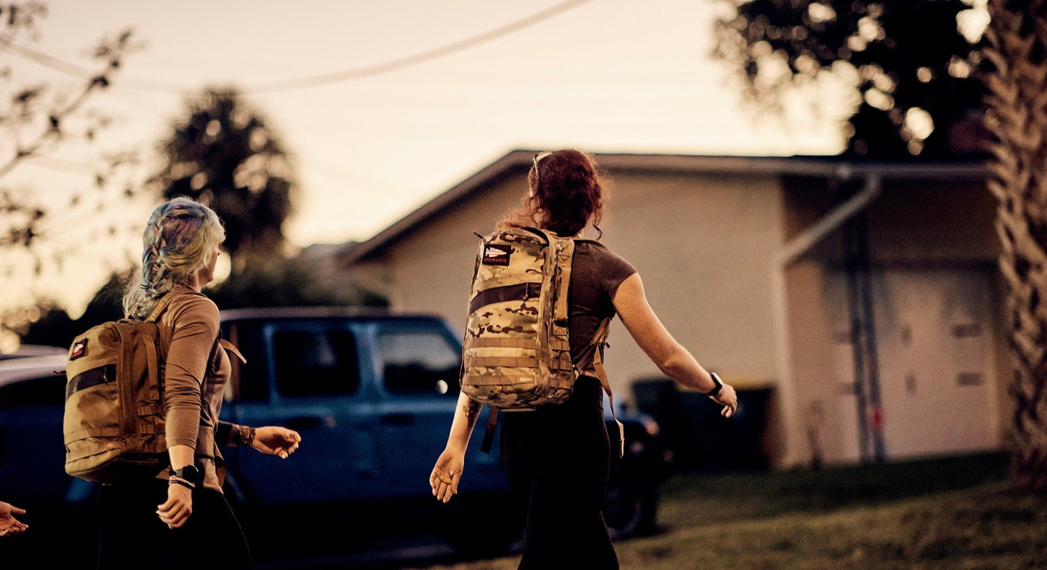 Two women wearing GORUCK backpacks walking outdoors near a house at dusk