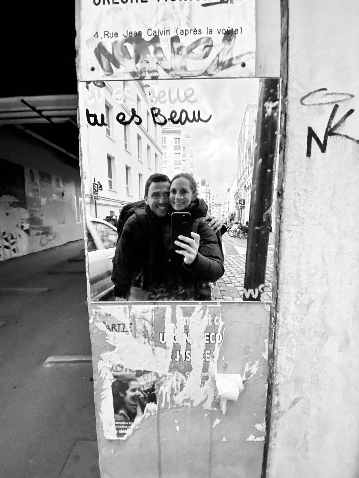 Smiling couple taking a selfie in a city street mirror, wearing outdoor gear, urban background.
