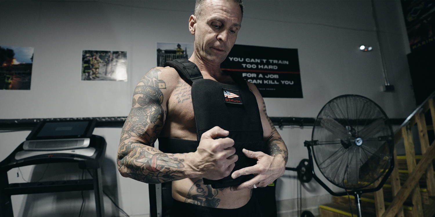 Tattooed man adjusting GORUCK weighted vest in gym, rucking gear and fitness equipment visible.