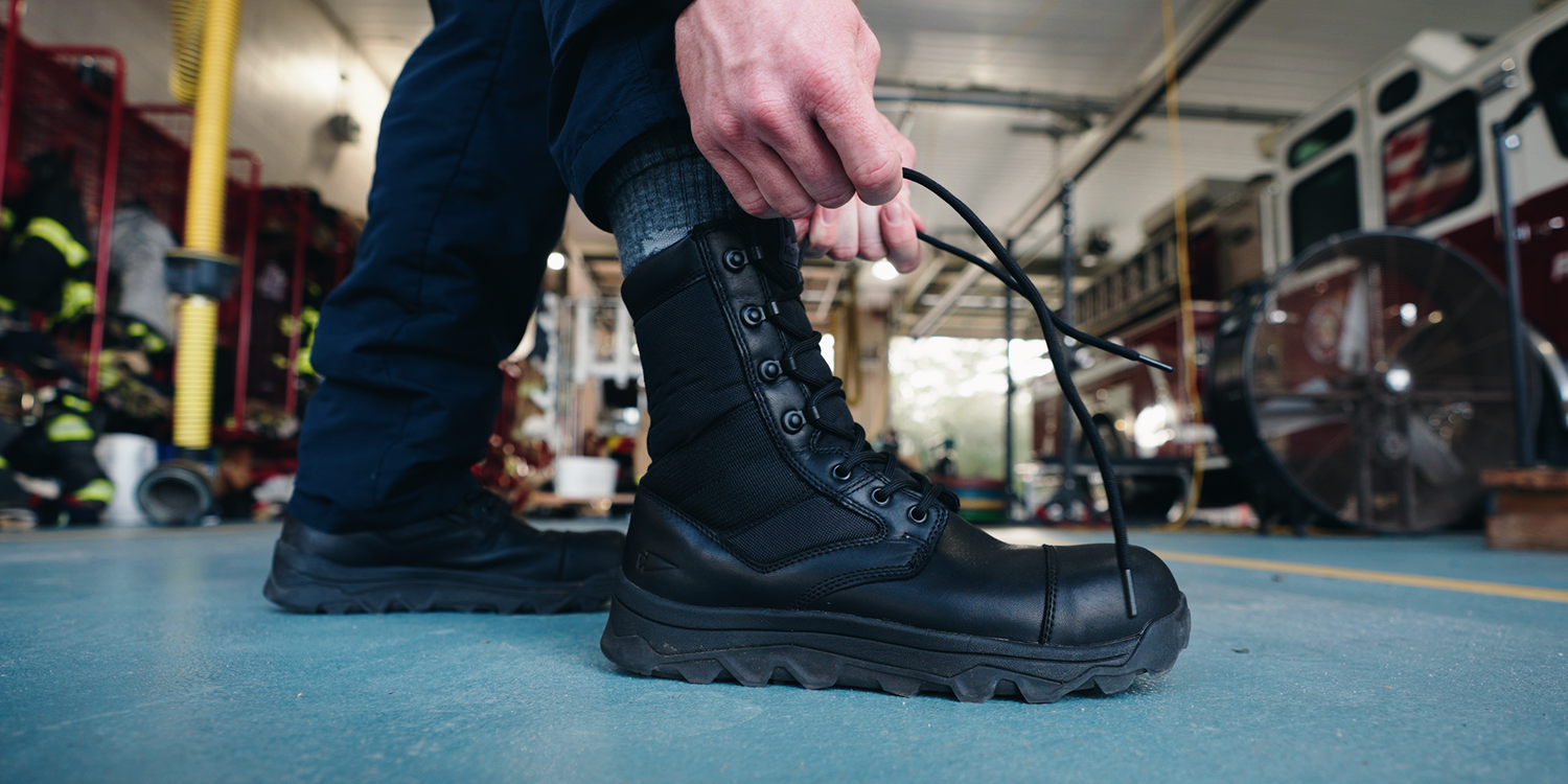 Person lacing up black goruck.momposite toe boot in fire station with gear and truck visible
