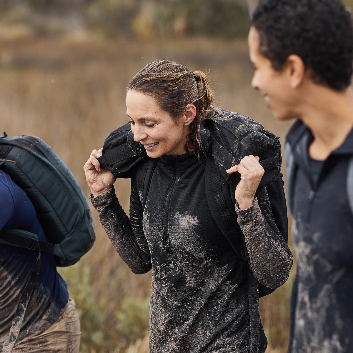 Smiling woman in black GORUCK gear with a muddy rucksack training outdoors with teammates