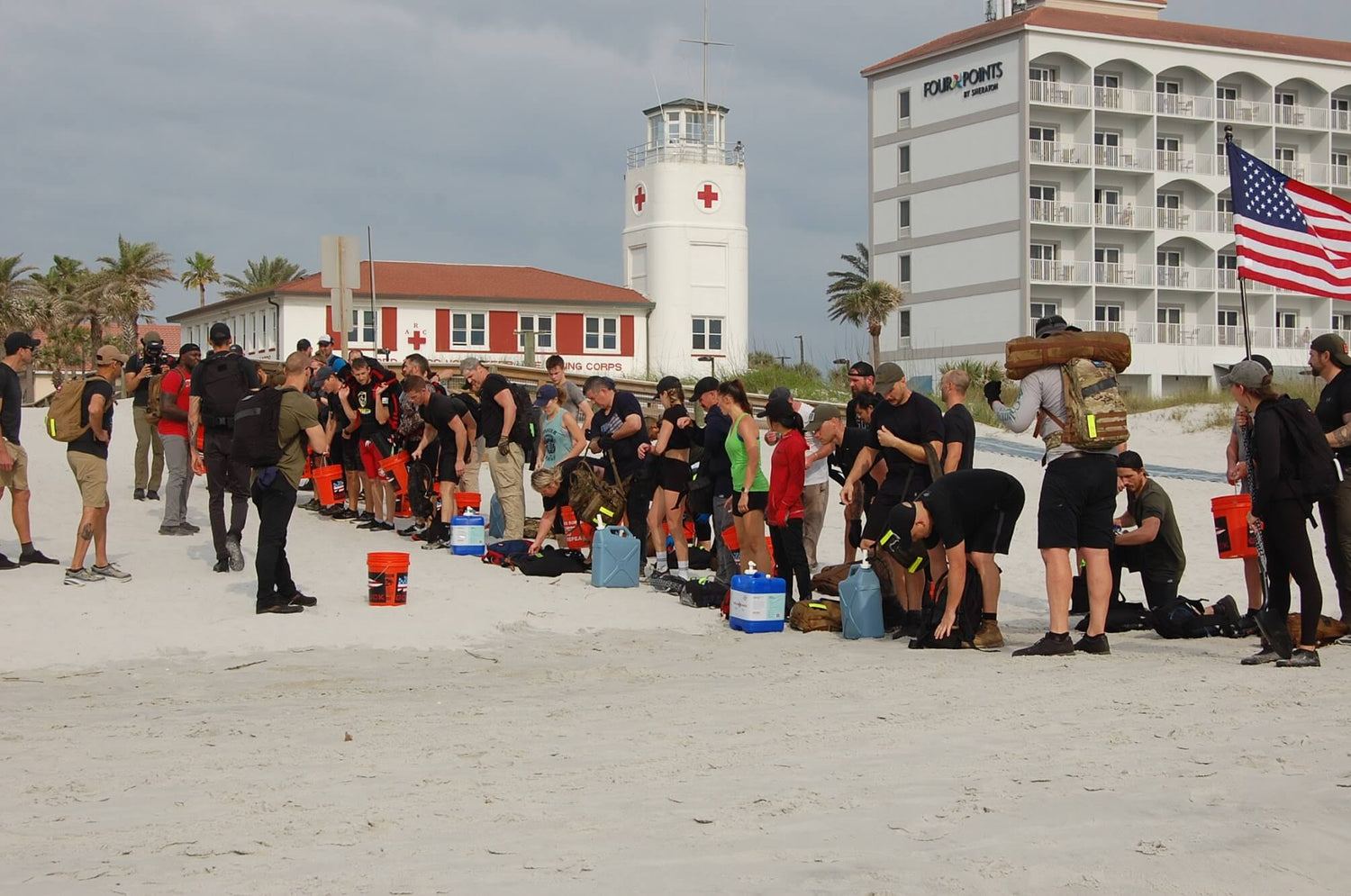 GORUCK rucking event participants on beach with gear, buckets, lighthouse, and American flag visible