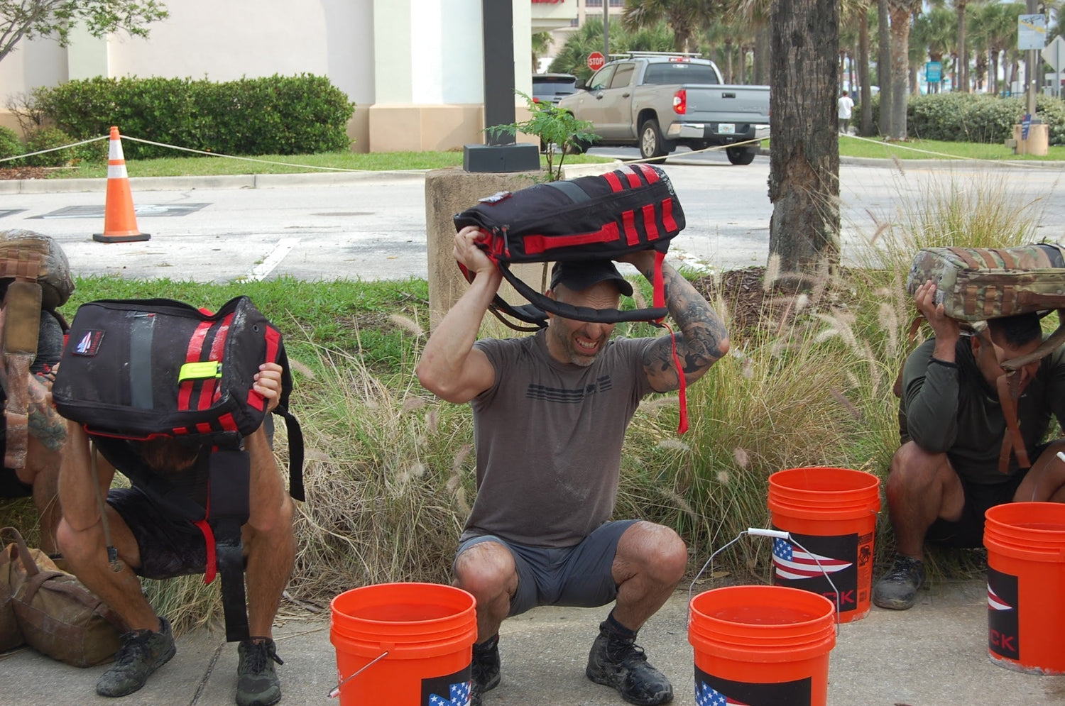 Three people squatting outdoors lifting GORUCK rucksacks, orange buckets in front, training event