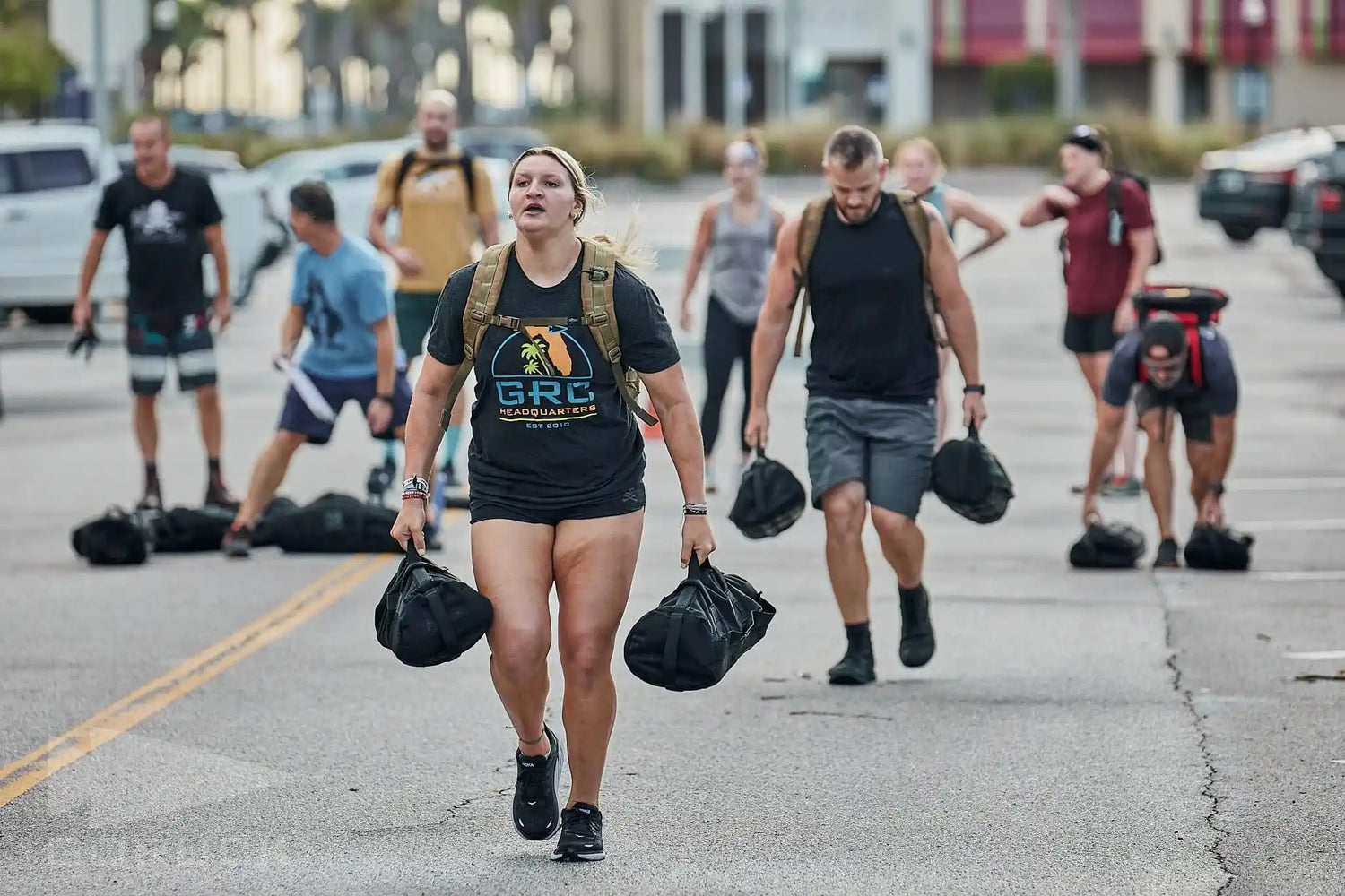 Group rucking outdoors with GORUCK gear, carrying heavy sandbags in a parking lot challenge.