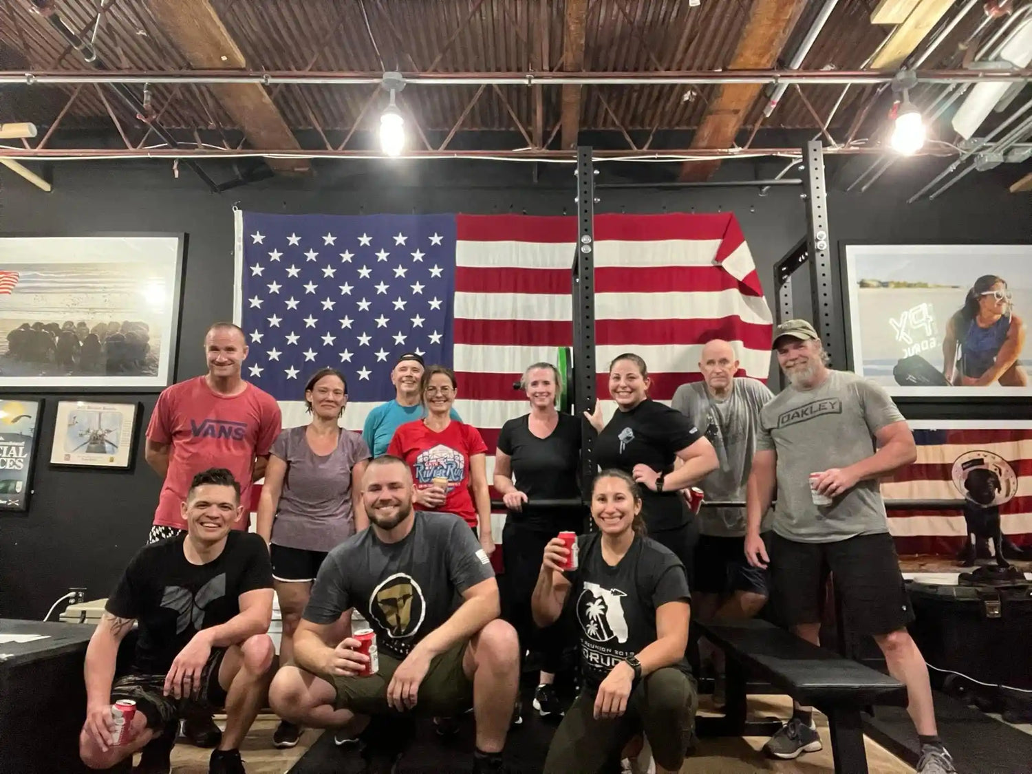 Group of people in athletic gear at GORUCK gym, American flag backdrop, smiling after rucking workout