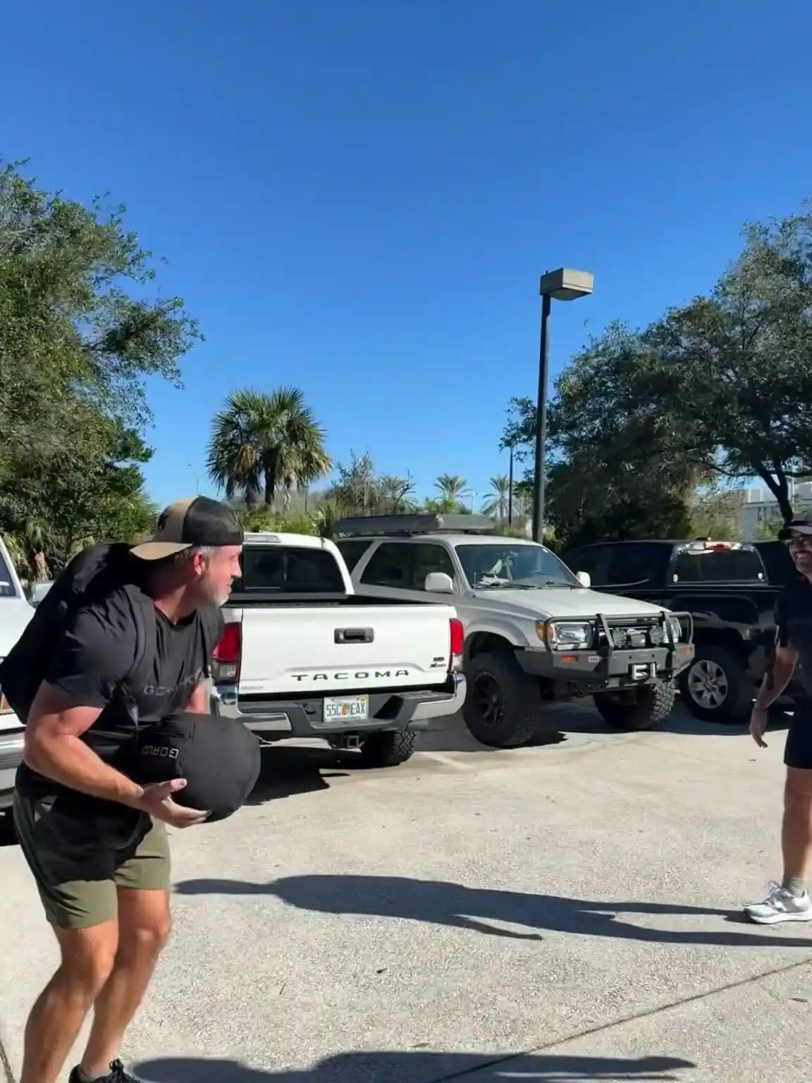 Man training outdoors with a GORUCK sandbag in a parking lot, wearing athletic gear