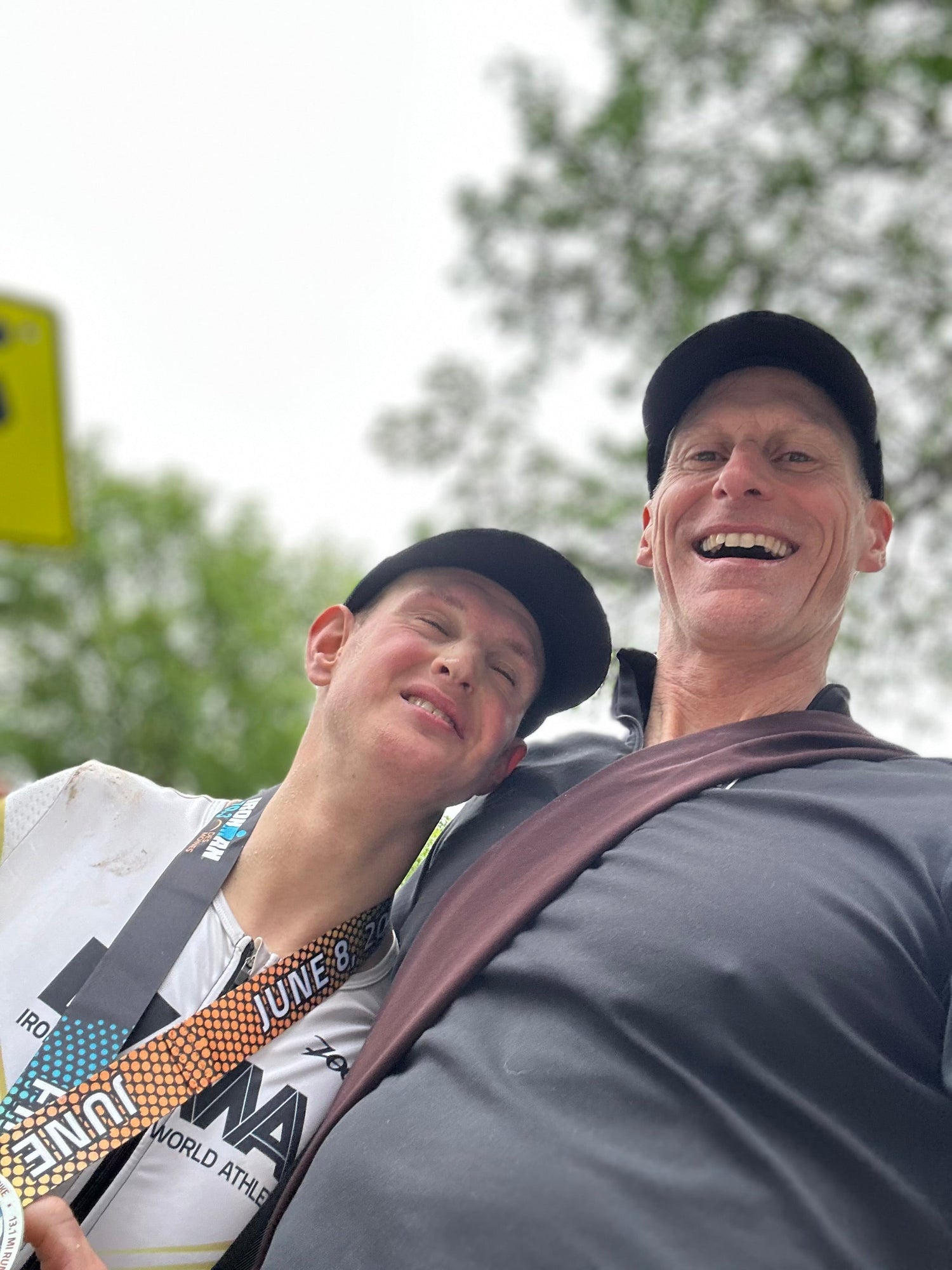 Two smiling men wearing hats, one with a race medal, outdoors under trees, goruck.mommunity spirit