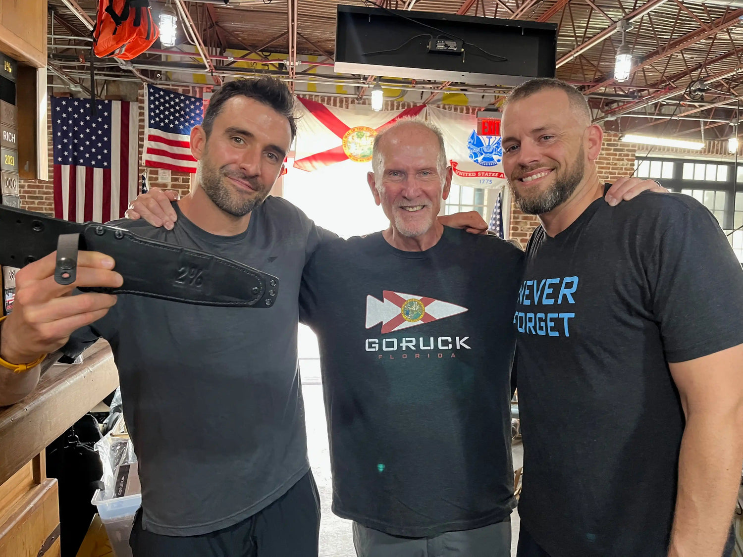 Three men at GORUCK event with American flags, smiling and wearing GORUCK branded shirts