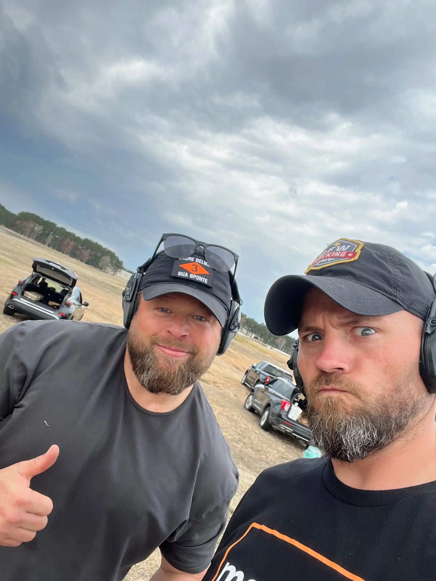 Two men in GORUCK clothing and caps outdoors at a rucking event, cars and cloudy sky behind them