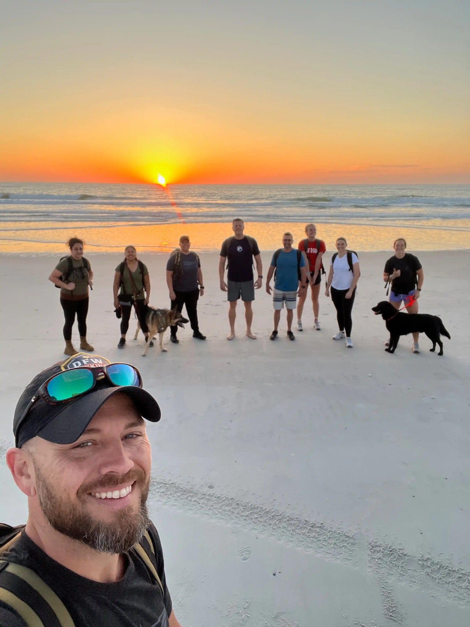 GORUCK group rucking on a sandy beach at sunrise, smiling with backpacks and dogs