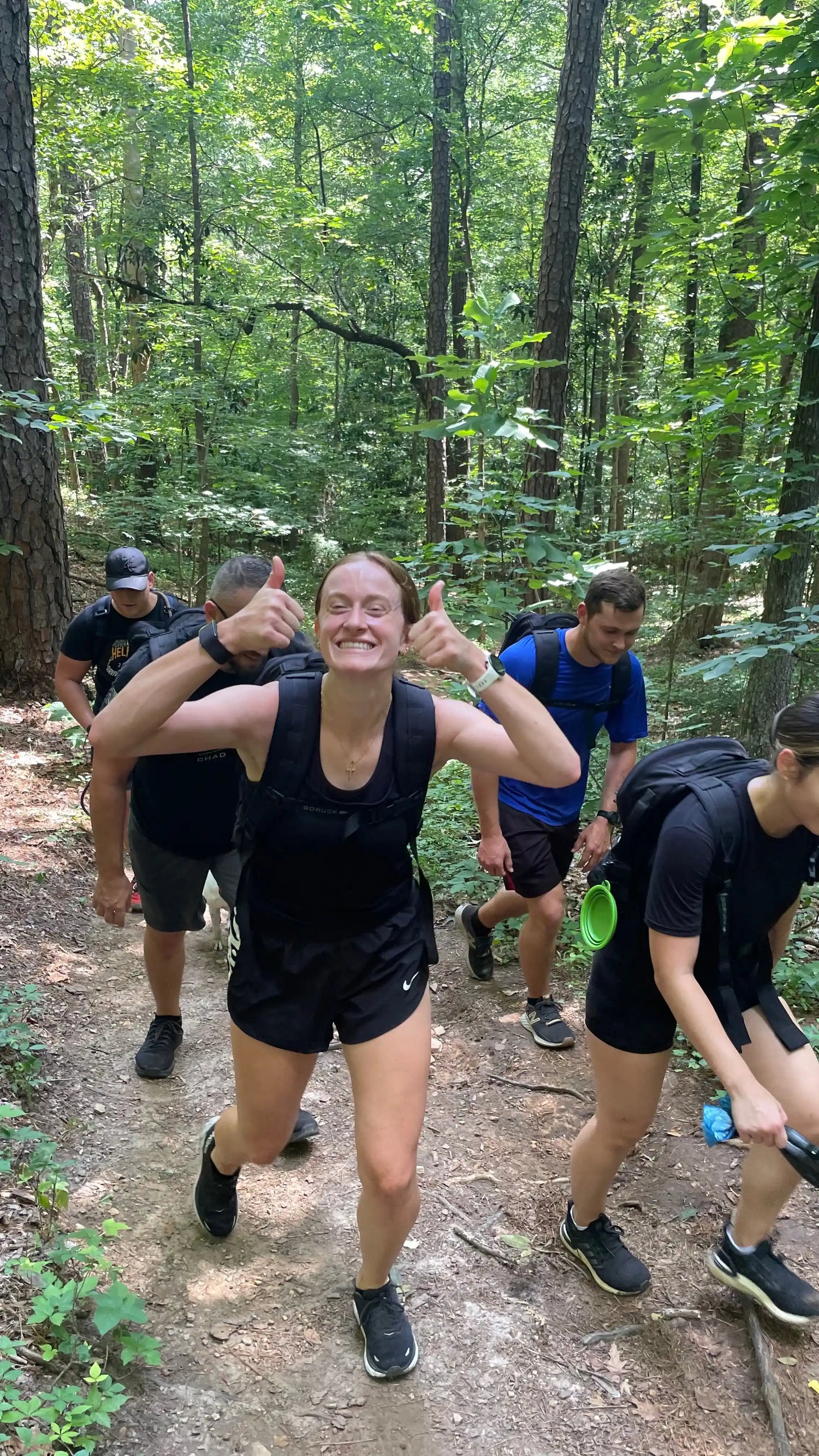 Group rucking on forest trail wearing GORUCK gear, smiling woman gives thumbs up