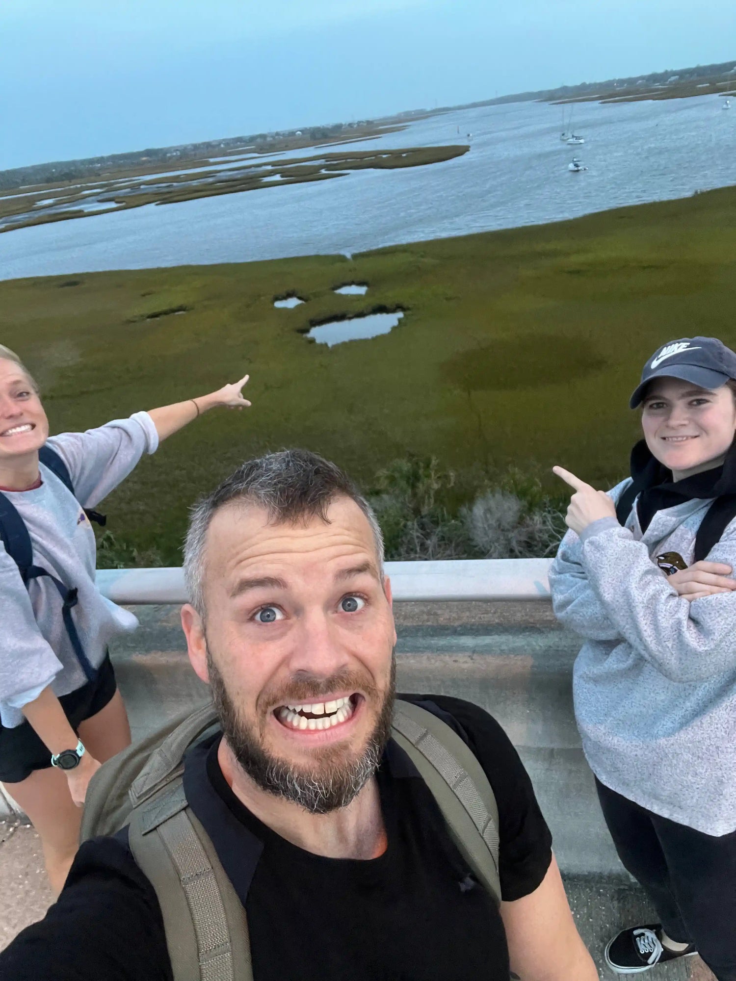Three people with rucksacks smiling on bridge, pointing at marshland with GORUCK gear in view