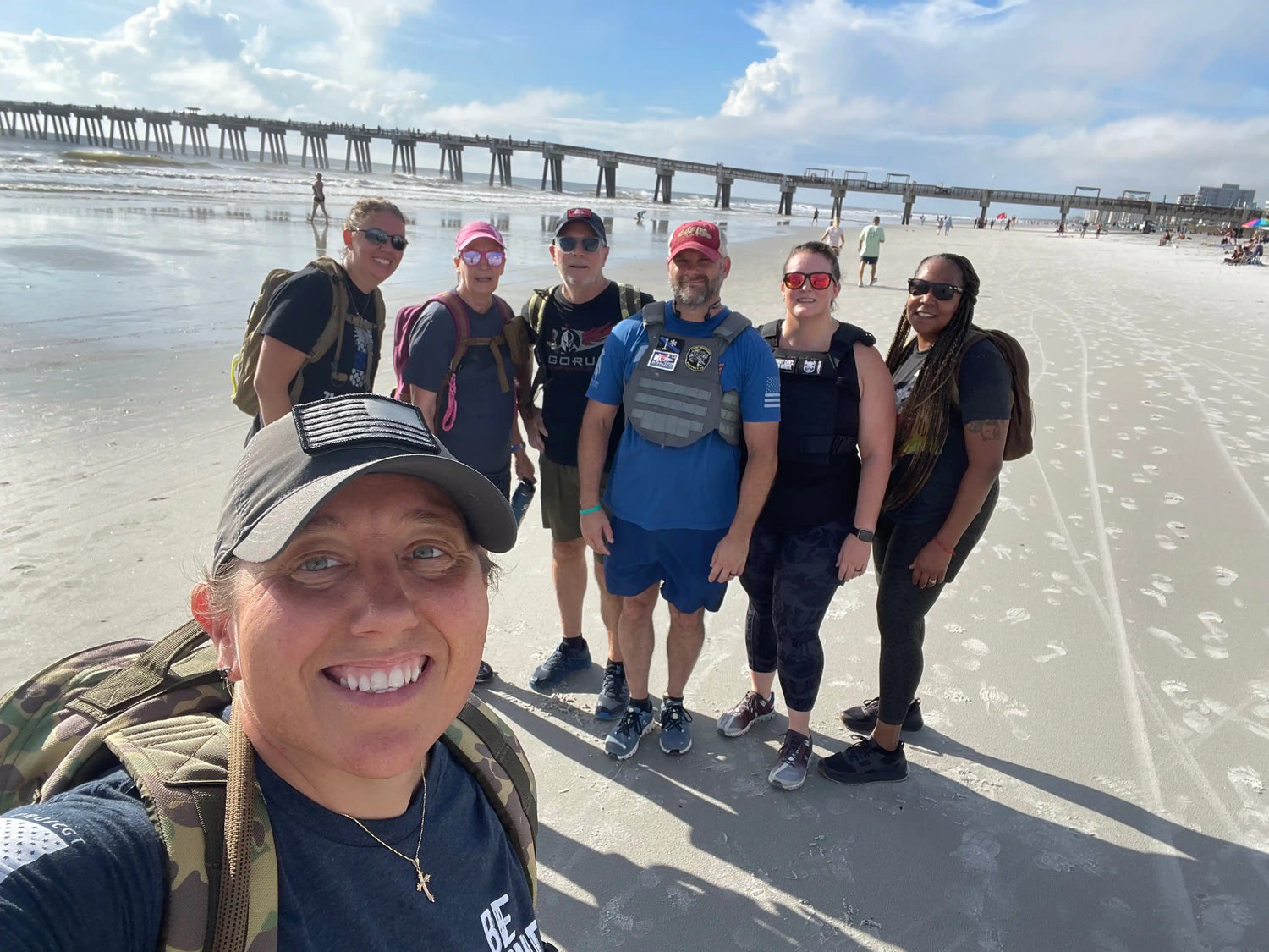 Group of people in GORUCK gear smiling on a sandy beach, pier in background, sunny day.