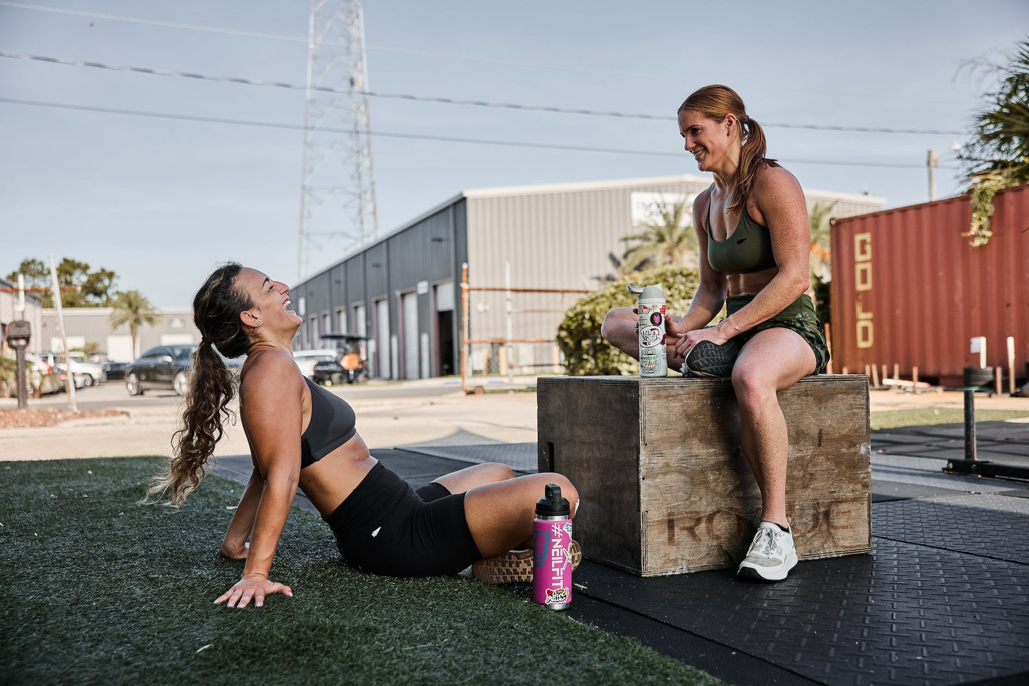 Two women in athletic gear relaxing and laughing after outdoor GORUCK workout by gym equipment
