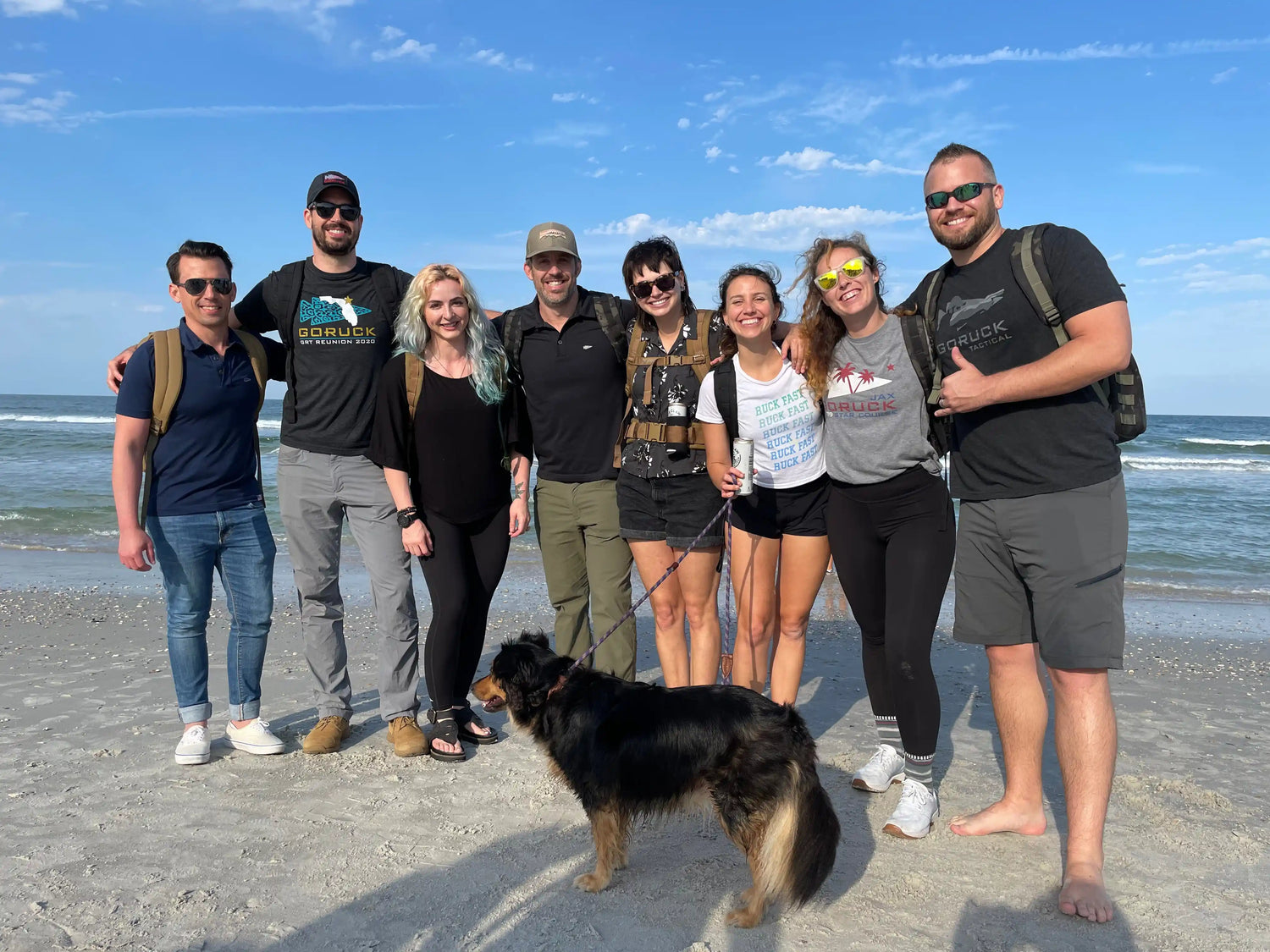 GORUCK team with backpacks and dog on sandy beach, group photo under blue sky