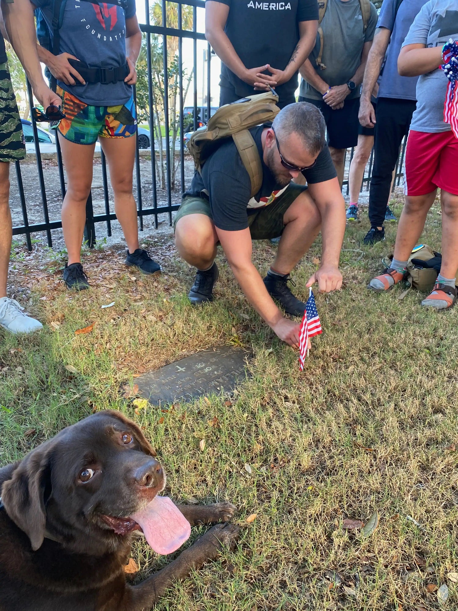 GORUCK group with backpacks places flag at memorial stone, brown dog in foreground