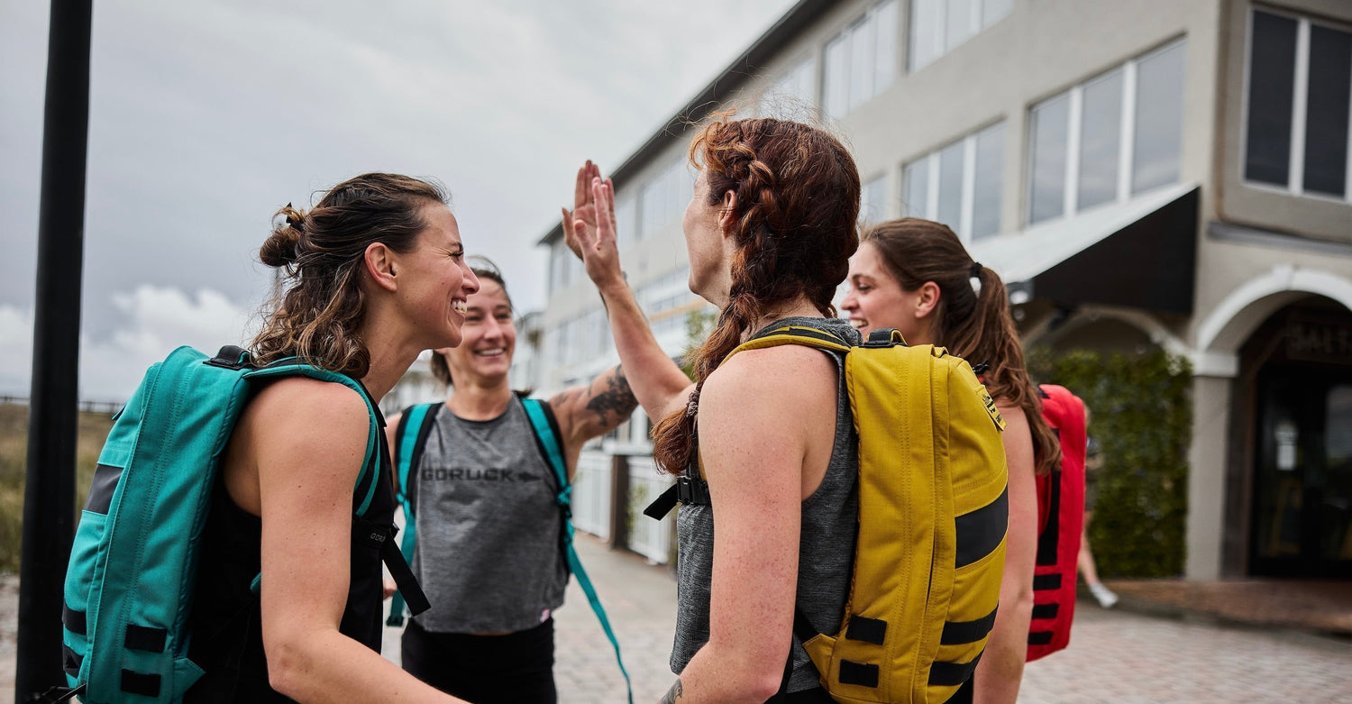 Group of women wearing GORUCK rucksacks outdoors, smiling and high-fiving