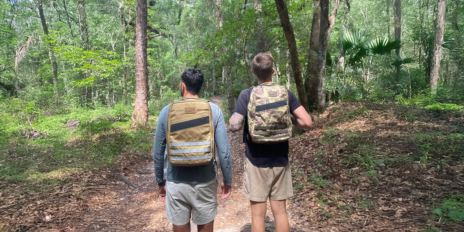 Two men hiking in the forest with GORUCK rucksacks, showcasing rugged outdoor gear.
