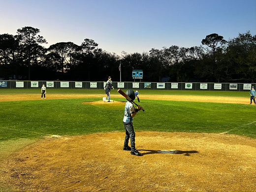 Youth baseball game at dusk, kid at bat, pitcher on mound, outdoor field, goruck.mommunity activity.
