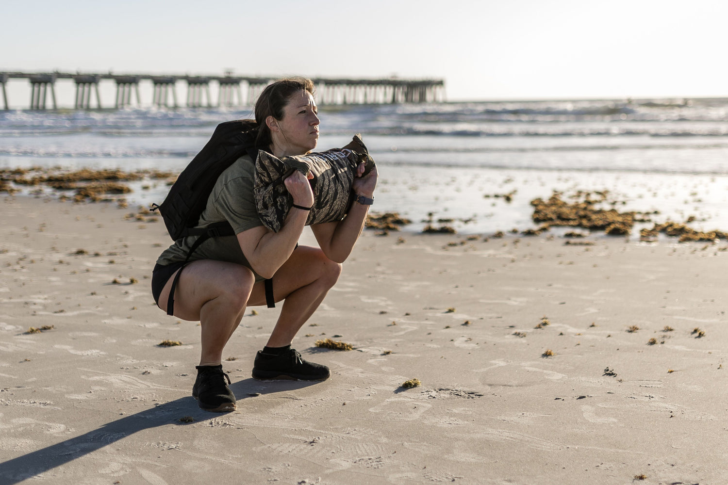 Woman rucking with sandbag on beach, GORUCK gear, ocean and pier in background