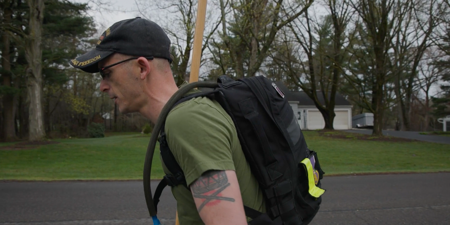 Man rucking outdoors with GORUCK backpack and hydration tube, wearing hat and sunglasses