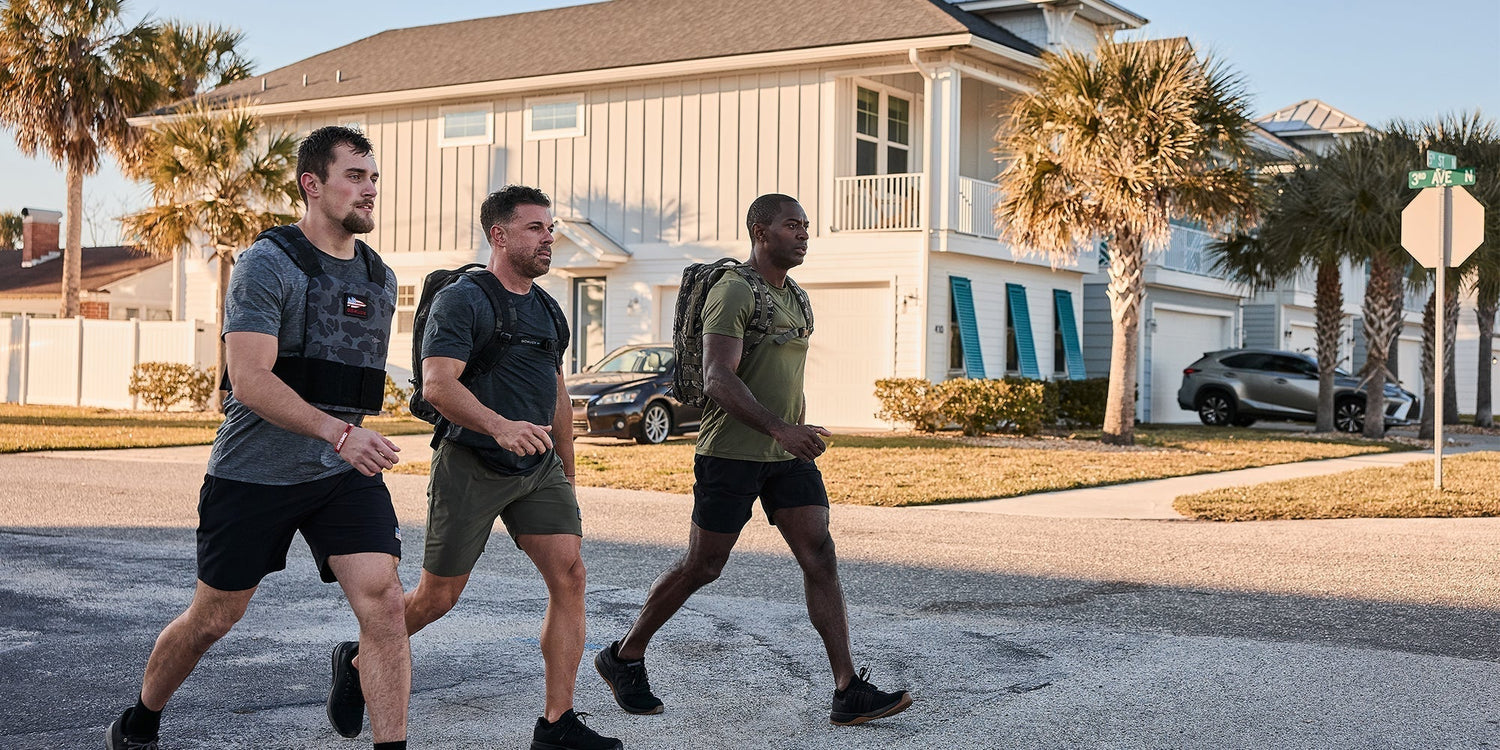 Three men rucking with GORUCK gear walk on a sunny suburban street near palm trees and houses.