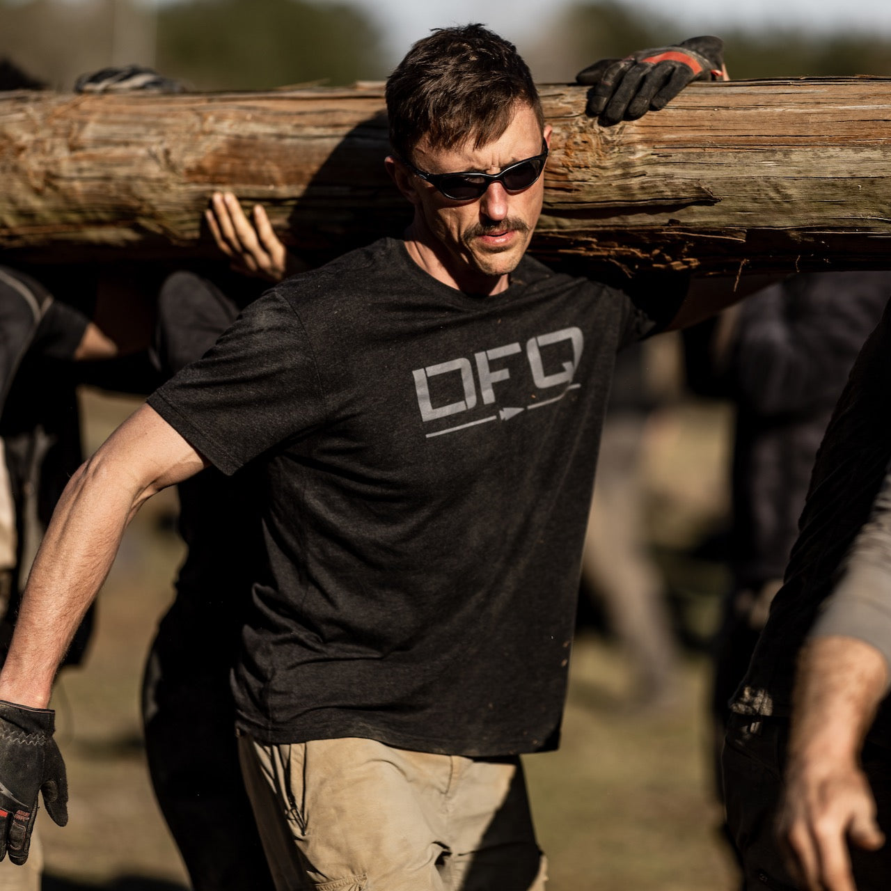 A man in sunglasses wearing a DFQ Tee - Tri-BlendX carries a large wooden log on his shoulder as he joins an outdoor group.