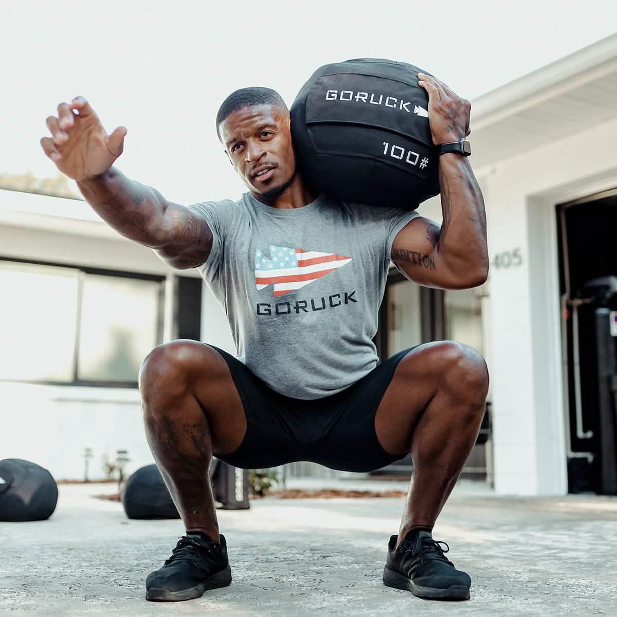 Man squatting outdoors with GORUCK 100 lb sandbag on shoulder, wearing GORUCK shirt