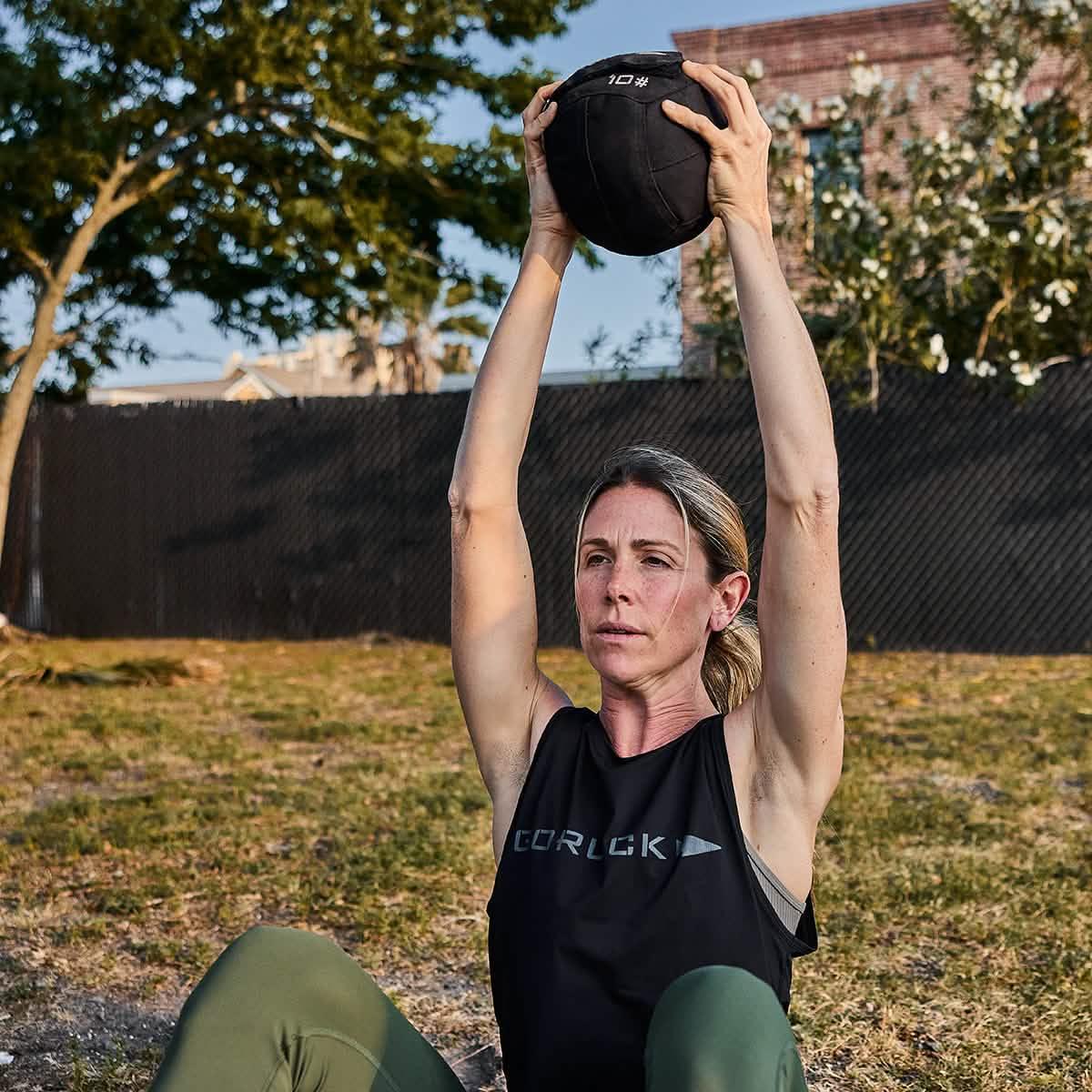 Woman training outdoors with GORUCK gear, holding 10lb medicine ball overhead