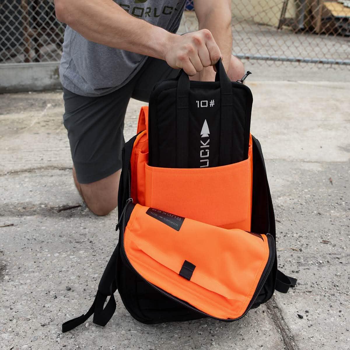 Person placing GORUCK 10lb black ruck plate into bright orange rucksack outdoors