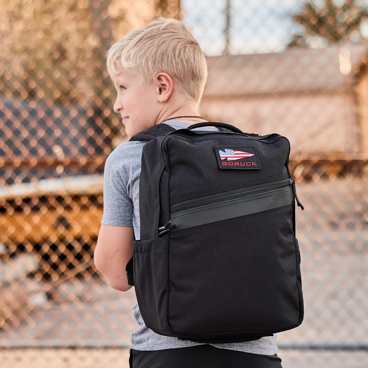 A young boy with short blonde hair is wearing a gray t-shirt and carrying the KR1 backpack from GORUCK. The black backpack, equipped with padded straps and an American flag patch, offers ample space for school supplies. He stands in front of a chain-link fence outdoors.
