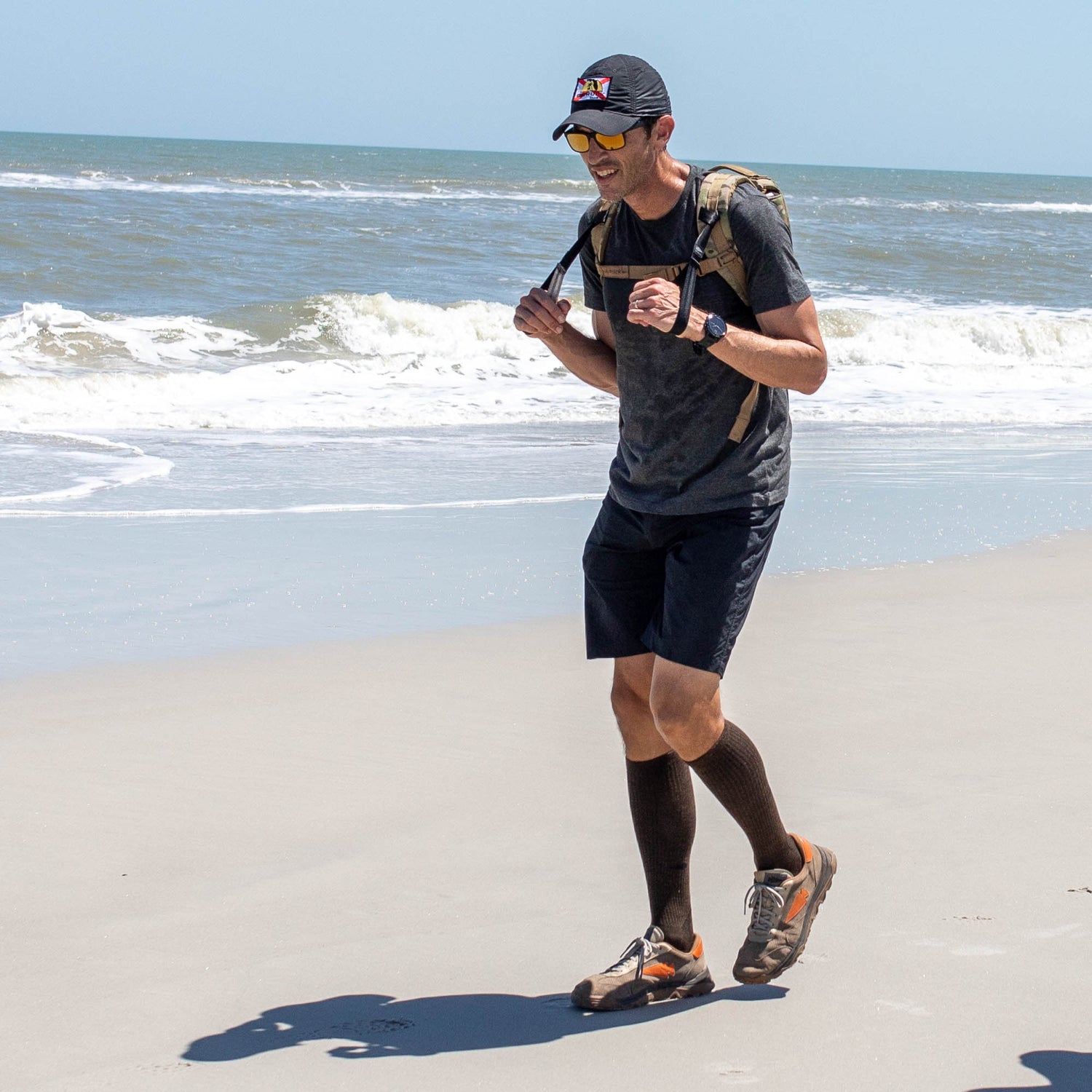 A man jogs on a sandy beach near the ocean, sporting a backpack, cap, sunglasses, athletic gear, and Merino Compression Socks for maximum comfort.