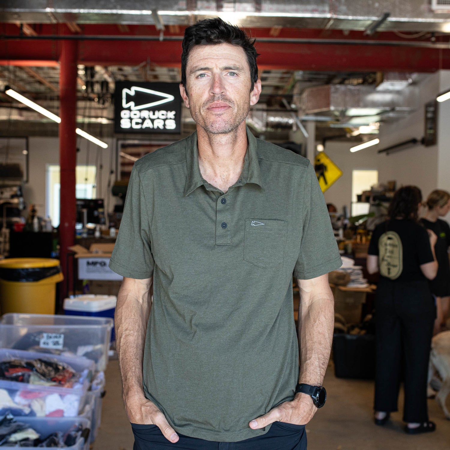 A man in an olive green Men's Polo with Pocket - Tri-BlendX stands in a busy workspace with a “GORUCK SCARS” sign in the background, highlighting the polo’s comfort and anti-stink features.
