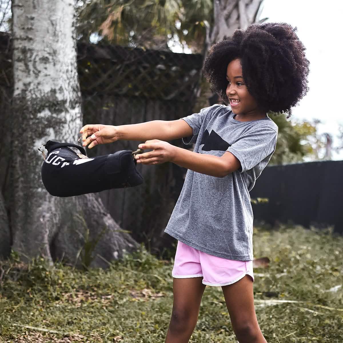 Smiling child using GORUCK training sandbag outdoors, wearing gray t-shirt and pink shorts