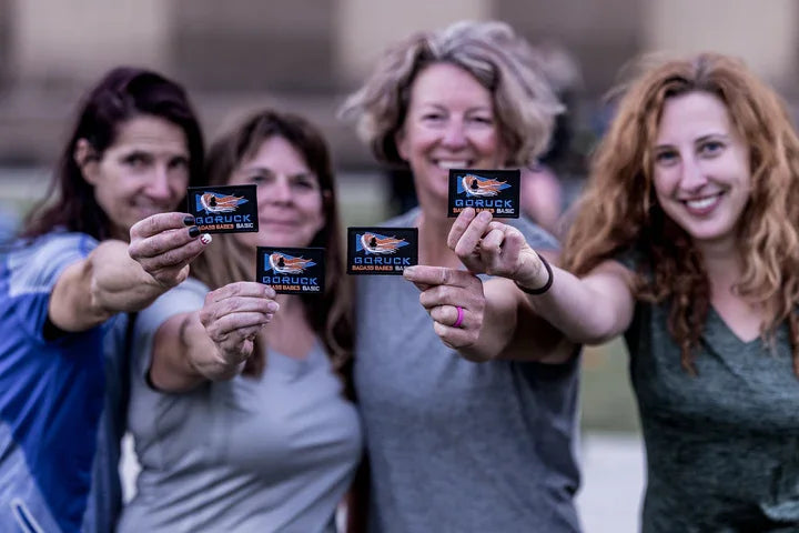 Four women smiling outdoors holding GORUCK patches, showcasing rucking gear community spirit