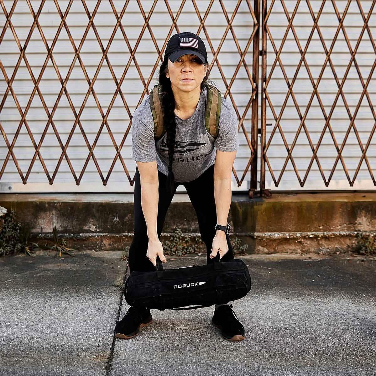 Woman rucking outdoors with GORUCK sandbag and rucksack, standing in front of metal gate.