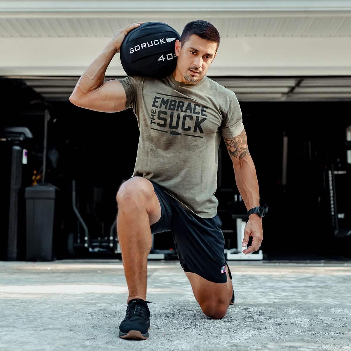 Man training with GORUCK weighted sandbag, wearing 'Embrace the Suck' shirt, outdoors