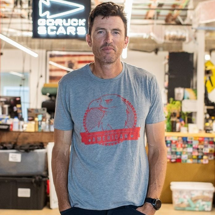 A man wearing the “100% American” Graphic T-Shirt stands in an industrial office space with colorful decor and an exposed ceiling.