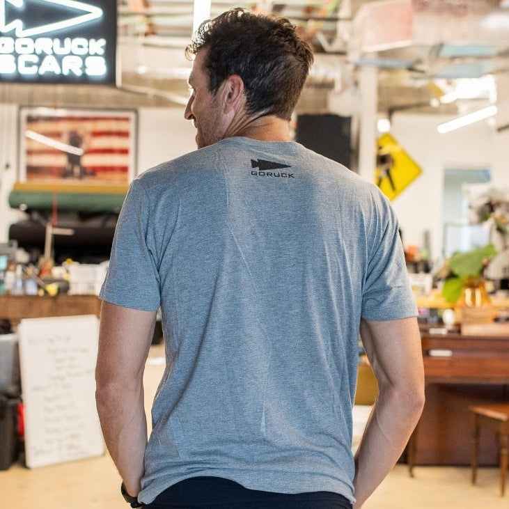 A man wearing the Graphic T-Shirt - Built in the USA stands indoors, facing away in a workspace filled with equipment and a GORUCK sign.