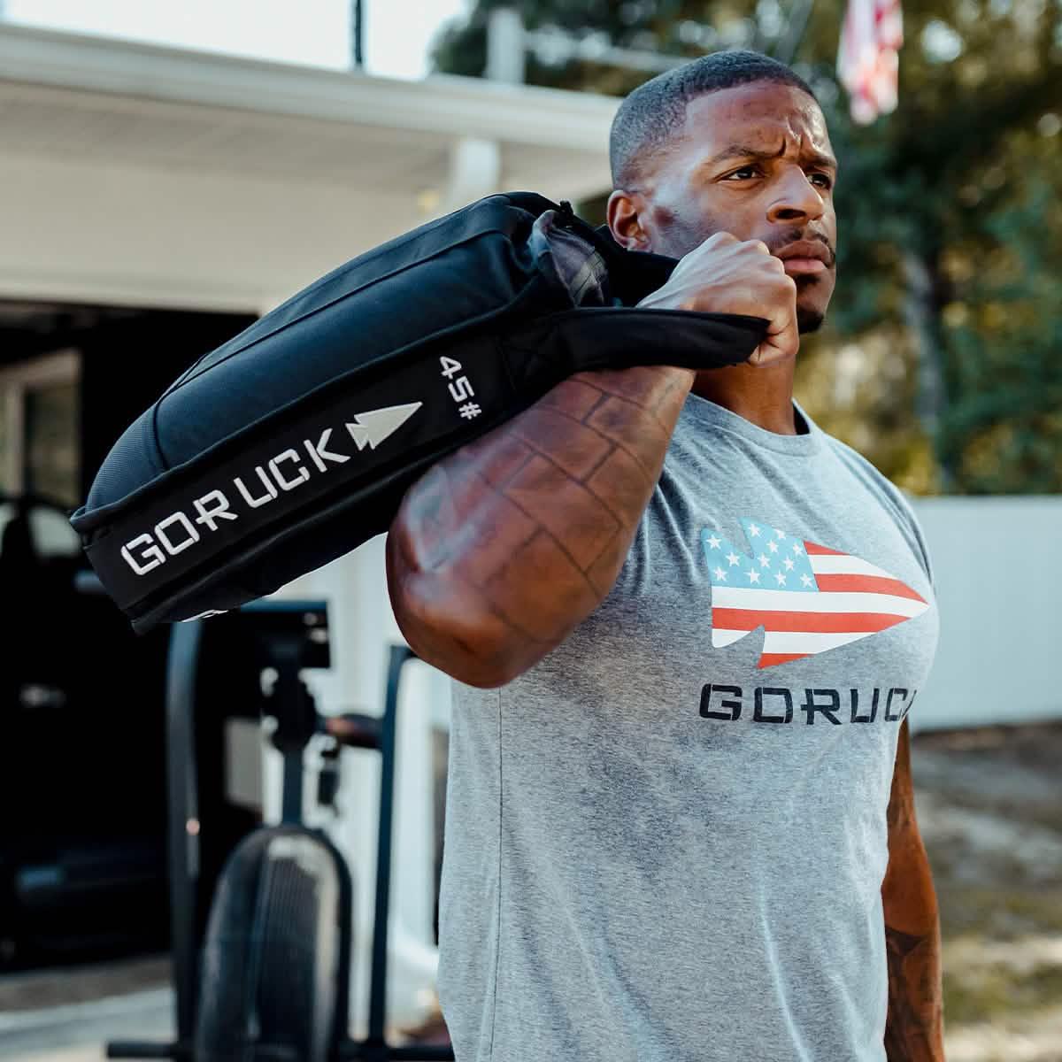 Man training outdoors with GORUCK 45 lb sandbag, wearing GORUCK logo shirt
