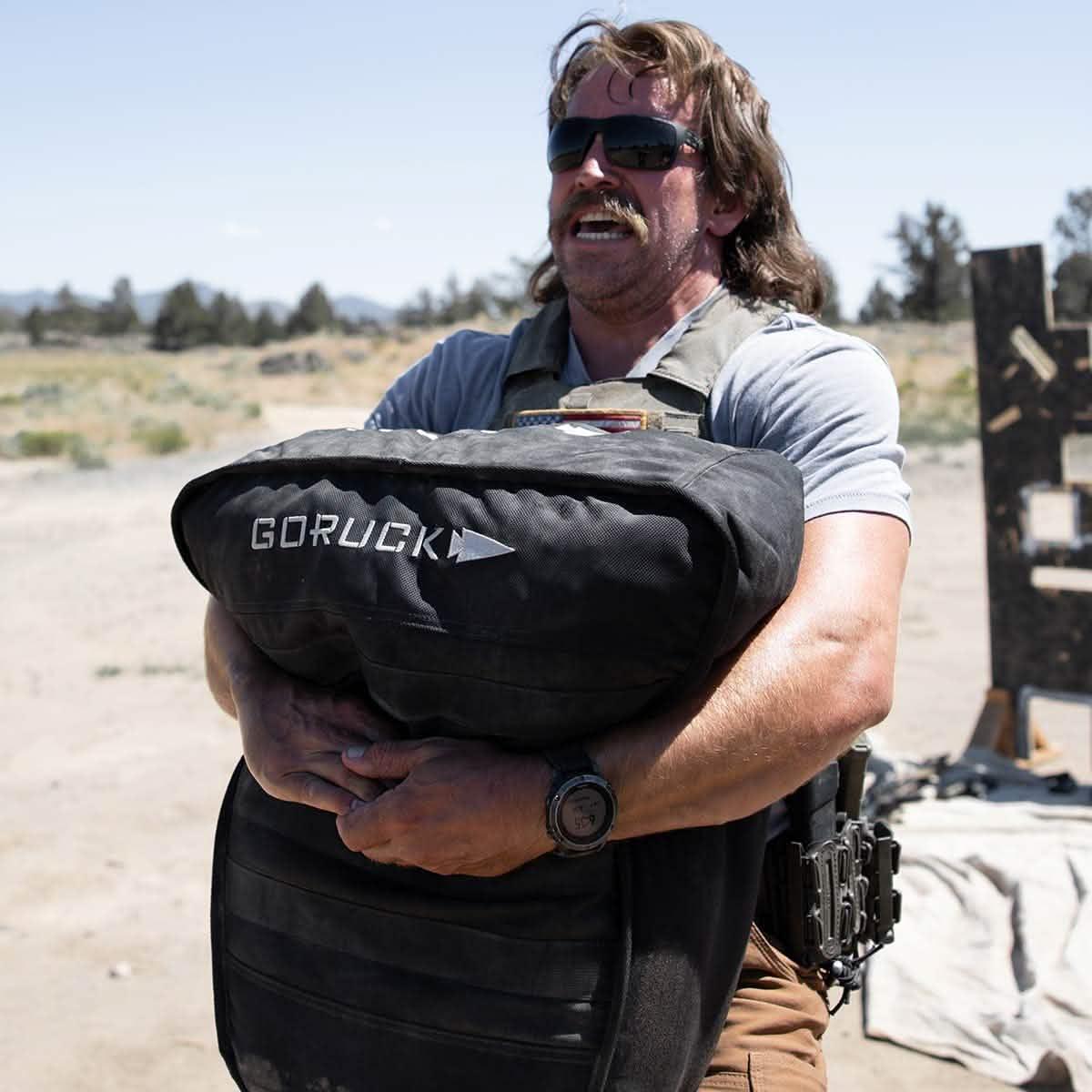 Man outdoors carrying GORUCK rucking sandbag, wearing tactical gear and sunglasses.