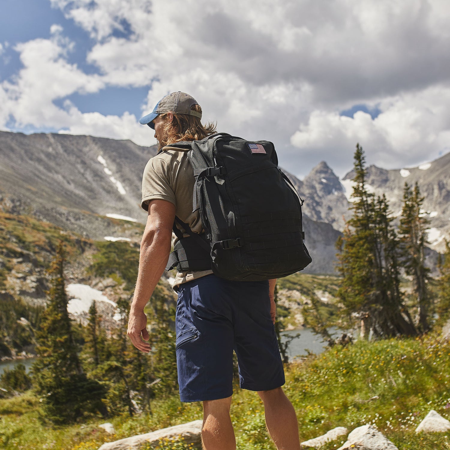 A man hikes through a mountain landscape, surrounded by trees and rocks, wearing Men’s Challenge Shorts - Lightweight ToughDry®, featuring technical design and cargo pockets under a cloudy sky.
