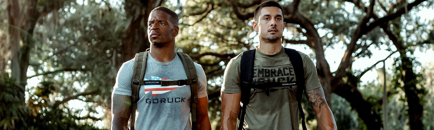 Two men with backpacks walk outdoors, wearing “GORUCK” shirts, surrounded by trees in daylight.