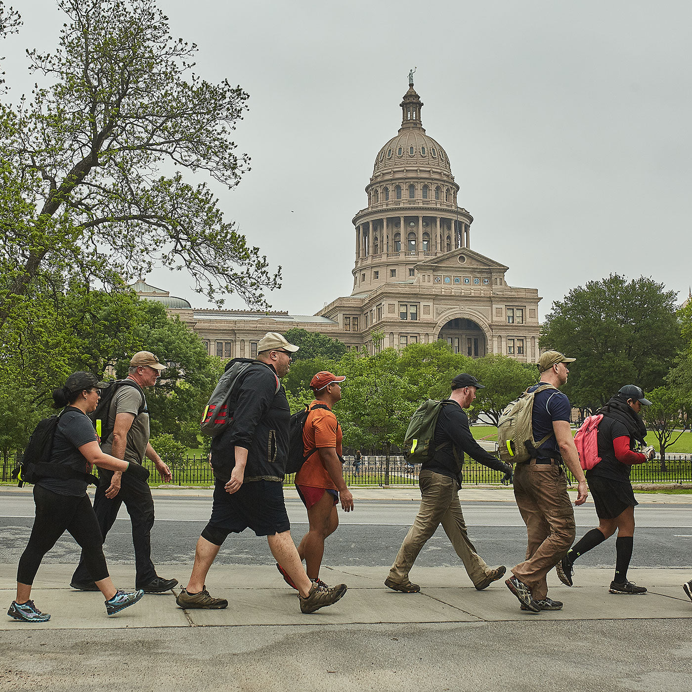 A group of people with backpacks walk past the Texas State Capitol building on a cloudy day.